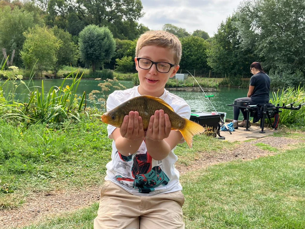 Young person holding crucian carp with lake and another person fishing in background. 